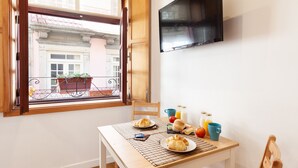 Ruokailutilassa on aamiainen katettuna: croissantteja, pullo appelsiinimehua ja kuppi. Seinälle on asennettu televisio, ja ikkunasta avautuu näkymä rakennuksiin ja parvekkeelle.