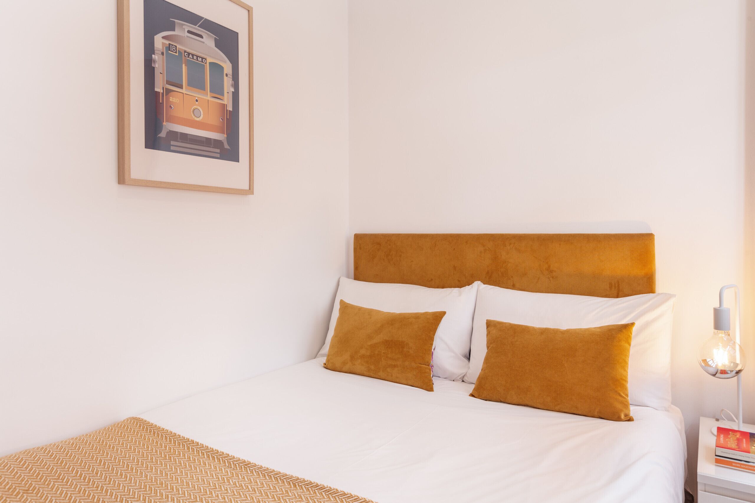 Una cama bien hecha con cabecera blanca y marrón, un cuadro en la pared y una mesita de noche con una lámpara y un libro.