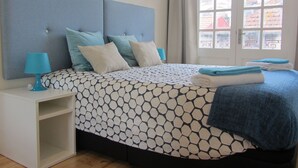 A neatly made bed with a patterned bedspread, a bedside table with a lamp, and a view of buildings through a window. - D&S - Crocodile House S. Bento Apartments IV (Oporto)