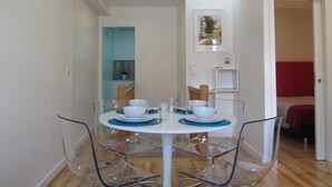 A dining area with a round table, clear chairs, and a blue placemat. There is a white wall with a framed picture and a door leading to another room. - D&S - Crocodile House S. Bento Apartments IV (Oporto)