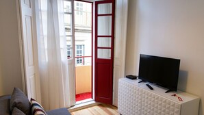 Ein Wohnzimmer mit einer Couch, einem Fernseher auf einer weißen Kommode und einer roten Tür, die zu einem Außenbereich führt.