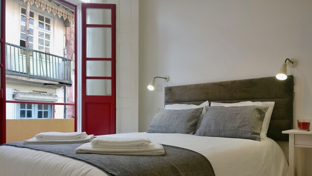 Ein ordentlich bezogenes Bett mit weißen Laken und einer grauen Tagesdecke, ein Nachttisch mit einer roten Tasse und ein Fenster mit roter Tür und Balkongeländer.