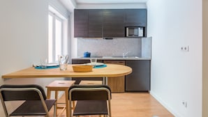 Een kleine keuken met donkere kastjes, een magnetron en een granieten aanrecht. Er staat een eettafel met stoelen en een klein raam zorgt voor natuurlijk licht.
