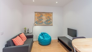 Een moderne woonkamer met een grijze bank, een blauwe pluchen zitbal, een houten eettafel en een flatscreen televisie.