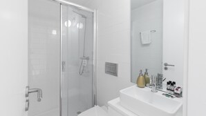 Un baño moderno con cabina de ducha de vidrio, azulejos blancos, un lavamanos con artículos de aseo y un espejo.