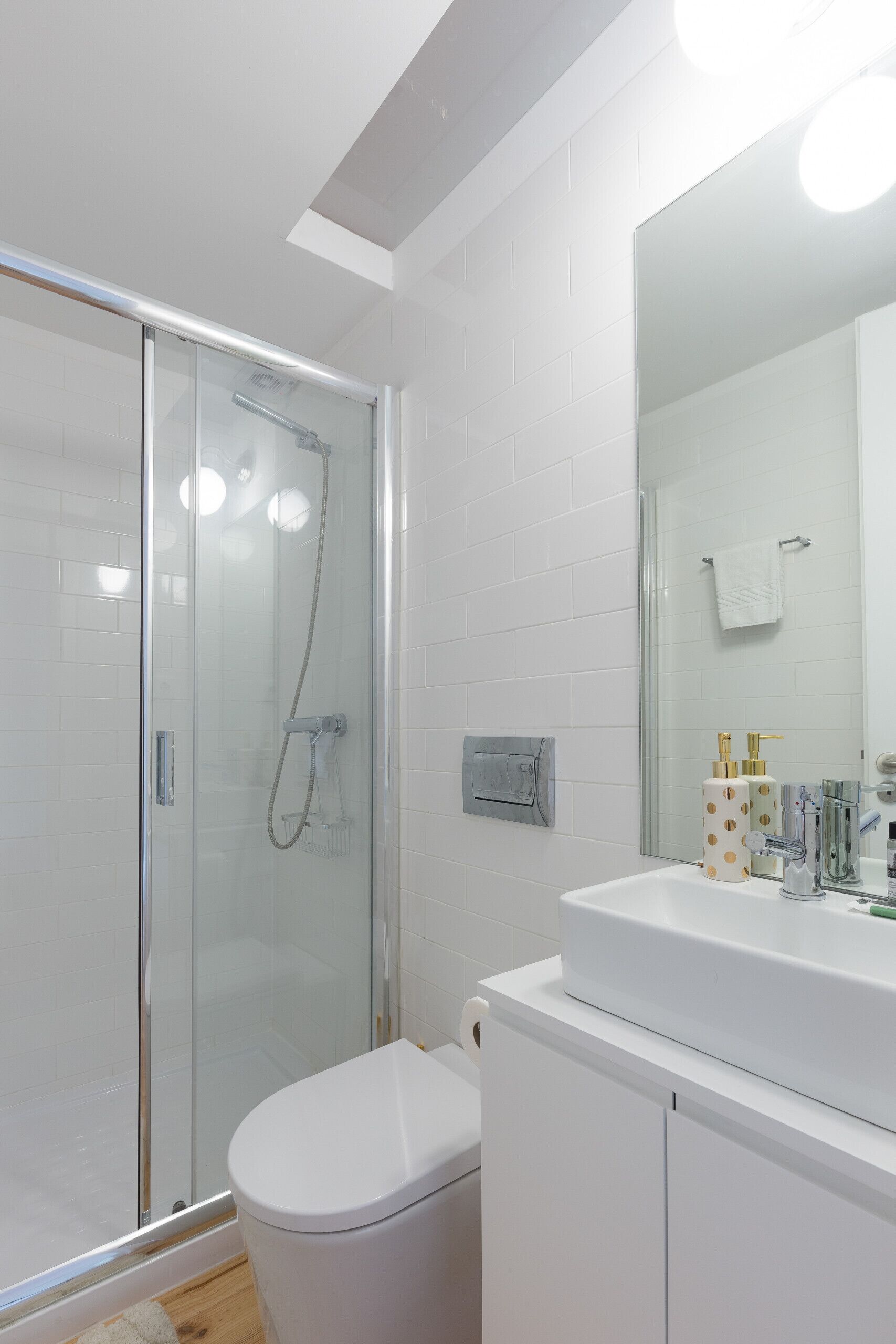 Un baño moderno con ducha acristalada, inodoro blanco y un espejo grande sobre un lavamanos blanco con artículos de aseo.