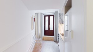 A hallway with a wooden floor, white walls, and a red door at the end. There are two clear chairs with gold legs and a small table.