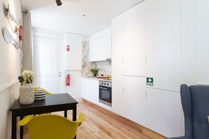 Une cuisine compacte avec des meubles blancs, un plan de travail noir et un plancher en bois. On y trouve une table à manger avec des chaises jaunes et un vase rempli de fleurs. Un extincteur est fixé au mur.