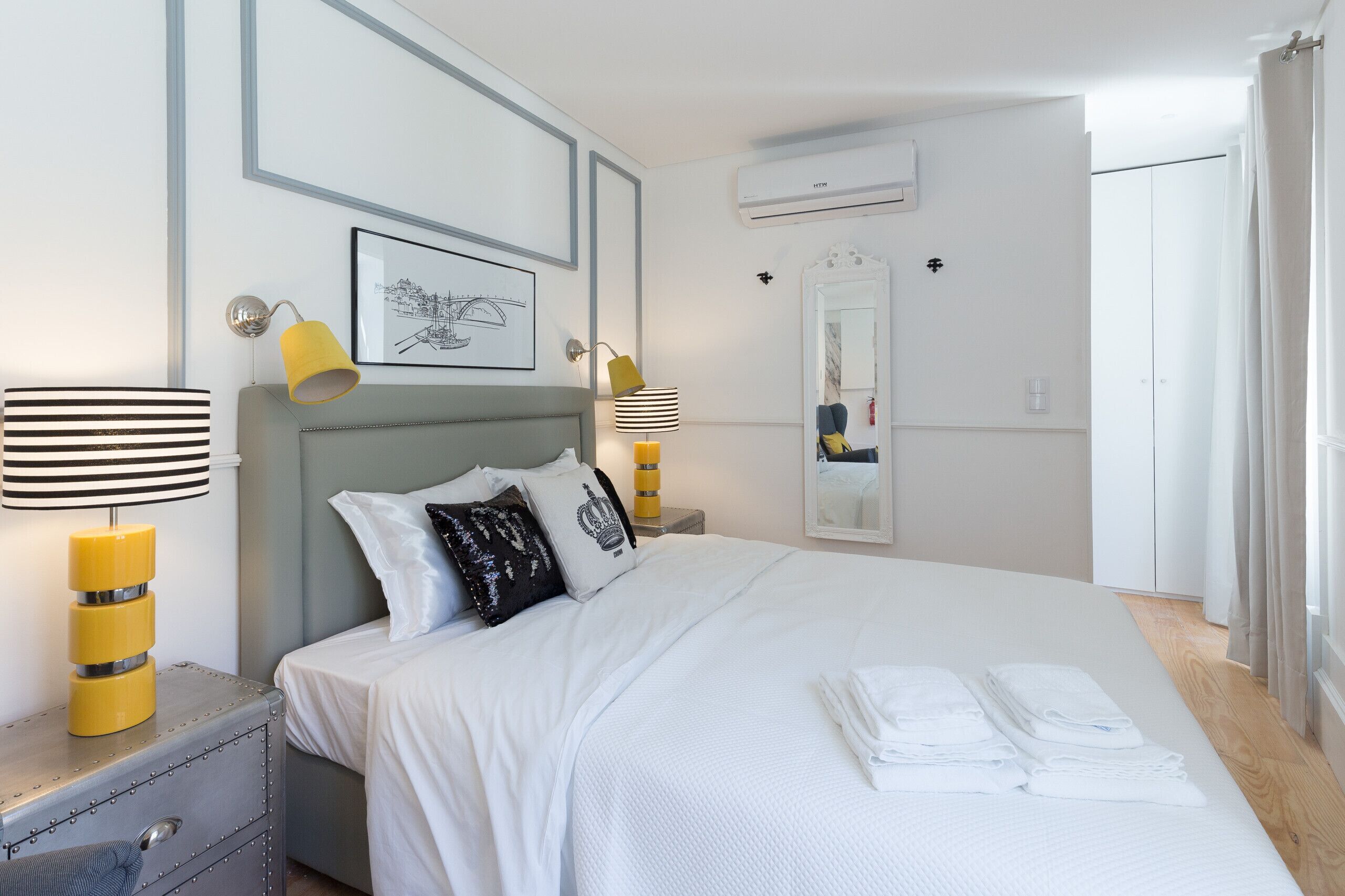 A neatly made bed with white linens, two bedside lamps, a mirror, and a suitcase on a nightstand.