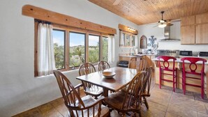 Uma área de refeições com mesa e cadeiras de madeira, uma cozinha com armários de madeira e grandes janelas com vista para o exterior.