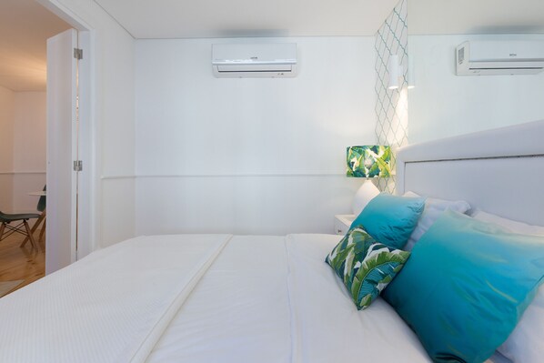 A hotel room with a neatly made bed, two wall-mounted air conditioners, a nightstand with a lamp, and a small table with chairs.