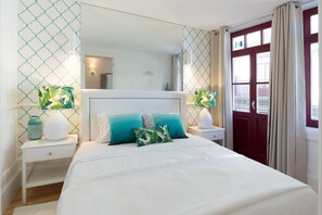 Ein Schlafzimmer mit einem weißen Bett, türkisfarbenen Kissen, einem weißen Nachttisch, einer weißen Lampe, einem Spiegel und einer roten Tür.