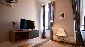 A modern living room with a flat-screen TV on a wooden console, a white chair, a small potted plant, and a view of the outdoors through large windows with curtains.