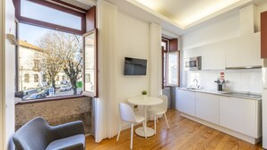 A modern kitchen with white cabinets, a microwave, and a sink. There is a dining area with a white table and chairs, and a gray chair. A TV is mounted on the wall above the dining area. - TRY OPORTO - Ponte do Infante (Oporto)