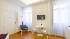 A modern hotel room with a wooden floor, a bed with a red blanket, a gray armchair, a white round table with two chairs, and a flat-screen TV mounted on the wall. - TRY OPORTO - Ponte do Infante (Oporto)