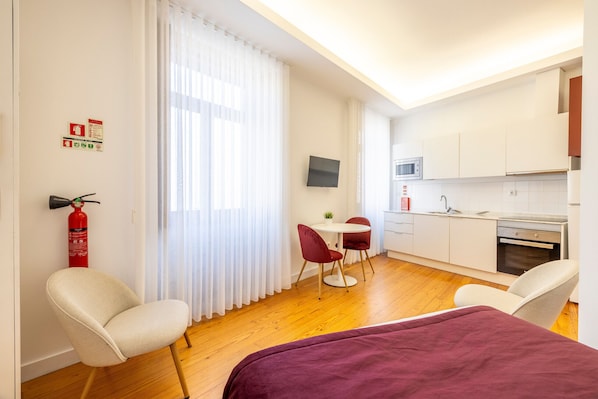 A compact hotel room with a bed, two chairs, a small table, a TV, a kitchenette, and a fire extinguisher.