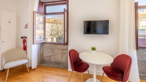 A room with a wooden floor, a white door, a red fire extinguisher, a TV on the wall, and a small table with two red chairs. - TRY OPORTO - Ponte Dom Luís (Oporto)