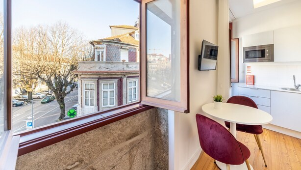 A modern kitchen with white cabinets and a microwave, a dining area with a white table and red chairs, and a large window with a view of a building and street. - TRY OPORTO - Ponte Dom Luís (Oporto)