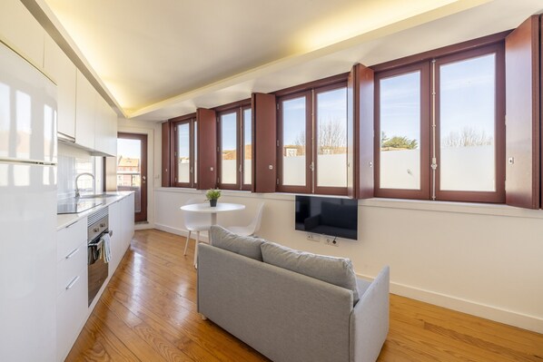 A modern kitchen with white cabinets, a built-in oven, and a dining area with a grey sofa and a small round table.