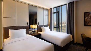 A hotel room with two beds, a large window with curtains, a desk with a lamp, and a chair.