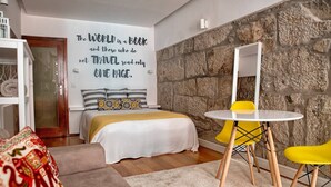 A modern bedroom with a stone wall, a bed with a patterned headboard, a round table with a yellow chair, and a large mirror. - Try Oporto Ribeira - Caves Port-Wine (Oporto)