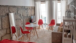 A modern hotel room with a stone accent wall, a bed with a red blanket, a sofa, a dining table with red chairs, and a large window with curtains. - Try Oporto Ribeira - Rio Douro (Oporto)