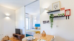 A modern room with a wooden dining table set for two, a television on a stand, a bed with a blue headboard, and a wall-mounted shelf with plants and decor. - D&S - Porto São Bento Apartments III (Oporto)