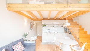 Un apartamento moderno estilo loft con cama en el altillo, un comedor con mesa y sillas redondas, una cocina con armarios blancos y una escalera con barandillas de madera.