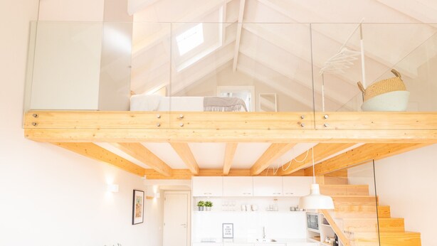 Un moderno espacio tipo loft con techo de vigas de madera, barandilla de vidrio y una zona de cocina en la parte inferior.