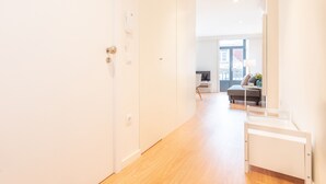 A bright, modern hallway with a wooden floor, leading to a room with a bed and a desk.