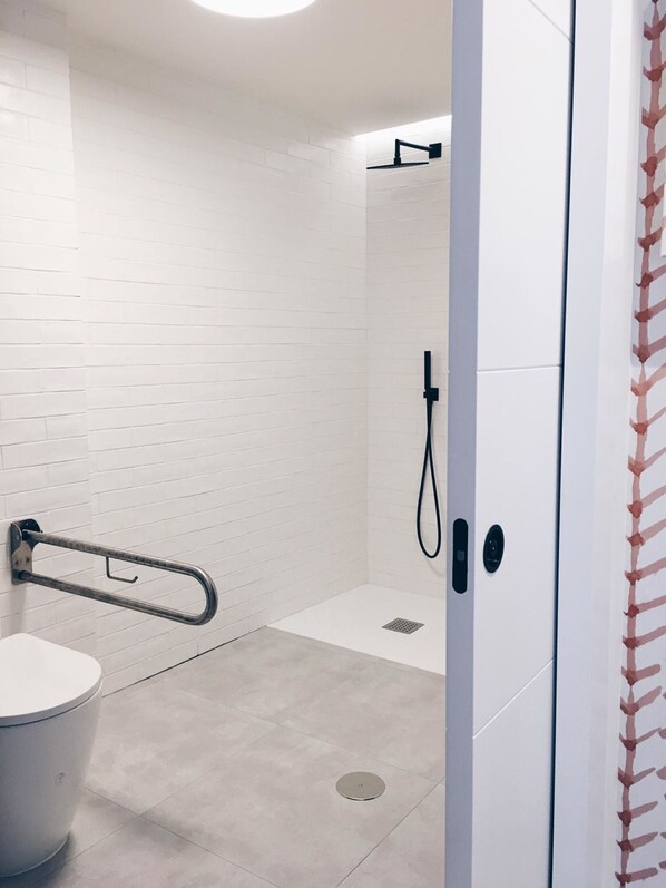 Une salle de bain moderne avec des carreaux de céramique blancs rectangulaires, une douche équipée d’un pommeau de douche à main et des toilettes dotées d’une barre d’appui de sécurité.