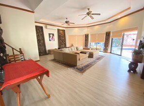 A spacious living room with a large sofa, a dining table with a red tablecloth, and a ceiling fan.