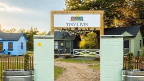 Un letrero de madera con la inscripción 'Cape Charles Tiny Livin', un camino de grava y una verja blanca que da a una casa.