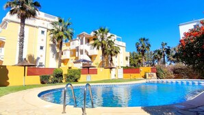 A swimming pool with a clear blue sky, a yellow building with balconies, and palm trees. - Apartament Playa Sol by DENIA COSTA (Denia)