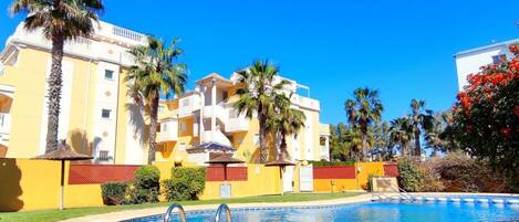 Una piscina con un cielo azul despejado, un edificio amarillo con balcones y palmeras.
