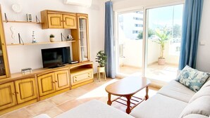 A living room with a sofa, a TV, wooden cabinets, a small table, and a balcony with a view. - Apartament Playa Sol by DENIA COSTA (Denia)