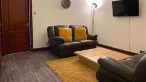 Un salon avec un canapé en cuir foncé, un tapis jaune, un téléviseur à écran plat et une horloge au mur.