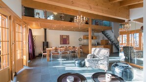 A spacious living area with a high ceiling, wooden beams, a chandelier, a dining area with chairs and tables, and a staircase leading to an upper level. - Waldbort Penthouse (Wengen)
