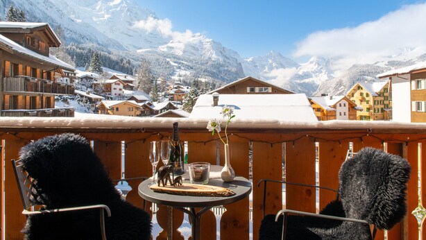A balcony with a table, two chairs, a vase with a flower, and a bottle of champagne. - Waldbort Penthouse (Wengen)