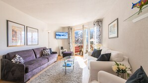 A living room with a purple sofa, a white sofa, a glass coffee table, and a TV on the wall.