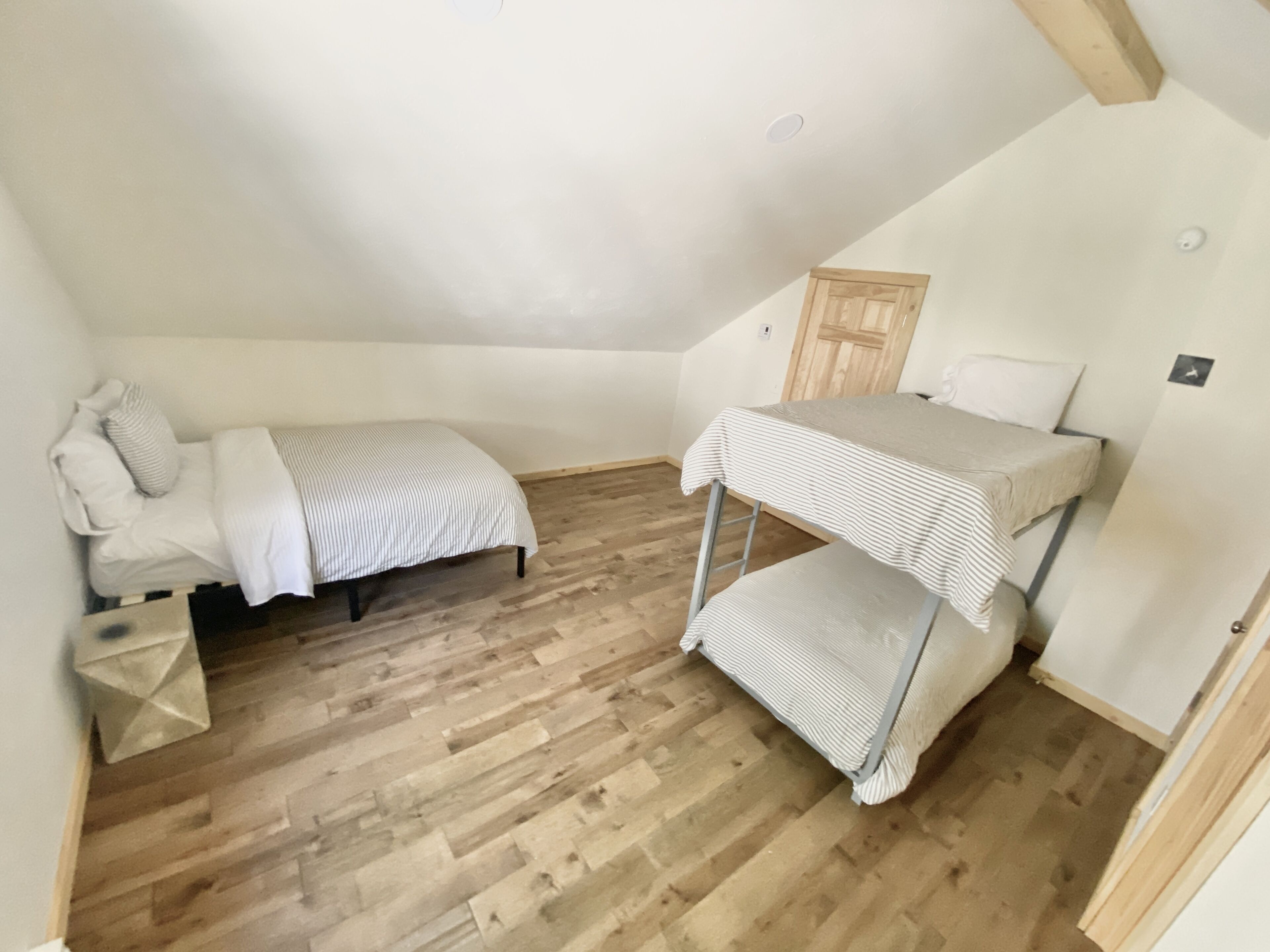 A small, single-room apartment with a sloped ceiling, wooden flooring, a bunk bed, a small table, and a door.