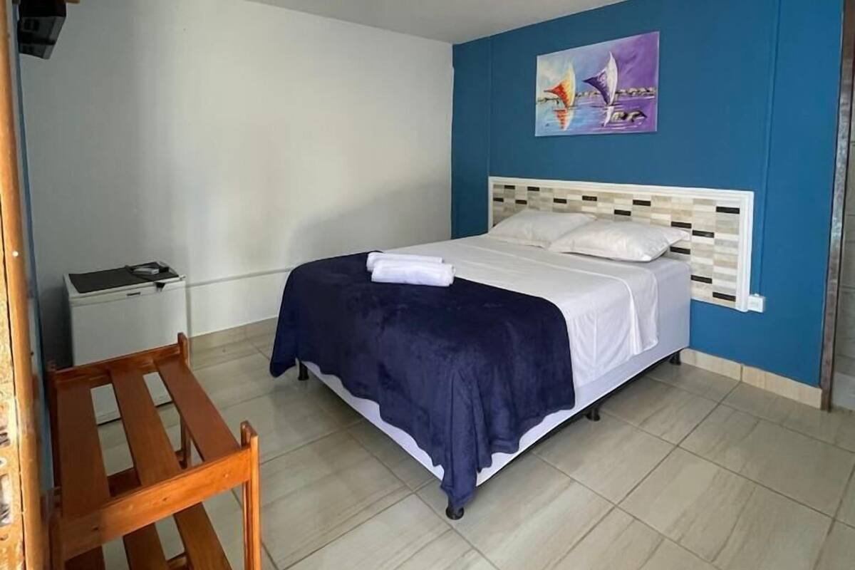A bedroom with a bed, a wooden chair, a small white cabinet, and a painting on the wall.