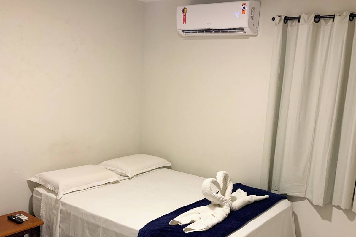 A hotel room with a bed, a bedside table, an air conditioning unit, and a curtain.