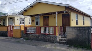 Uma casa amarela de dois andares com varanda, um portão vermelho e um muro de pedra.