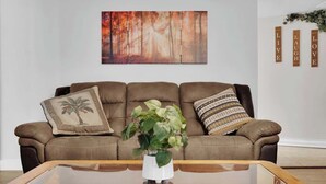 Een bruine leren bank met decoratieve kussens, een glazen bijzettafel met een plant en een schilderij met een boslandschap erboven.