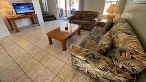 Ein Wohnzimmer mit Sofa, Couchtisch, Fernseher und einer Schiebetür zu einem Außenbereich.
