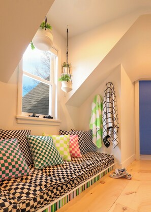 Une chambre à coucher chaleureuse, aménagée en style « loft », avec un canapé à carreaux, des plantes suspendues et une vue sur le ciel par une fenêtre.