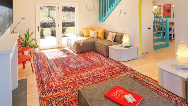 A modern living room with a colorful rug, a sofa with cushions, a coffee table with a book, and a staircase with a blue railing. - ⟰ DELIGHTFULLY DESIGNED (Boyne City)