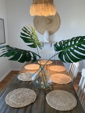A dining table with a clear glass vase, surrounded by woven placemats, and large green monstera leaves. - Happy House, minutes to the Beach! (Galveston)