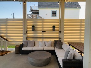 A covered outdoor seating area with a sofa and ottoman, a wooden table, and a railing. - Happy House, minutes to the Beach! (Galveston)